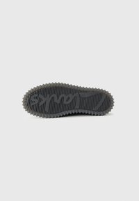 Black rubber shoe sole with textured grooves and circular accents. Features embossed logo and raised edges for grip.