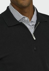 Schwarzer Strickpullover mit Reißverschlusskragen über einem weiß-blauen gestreiften Hemd mit Knöpfen. Verfügt über einen strukturierten Stoff und einen metallic Reißverschluss-Zipper.