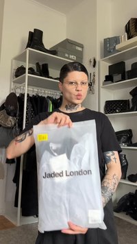Išsami, permatomoje maišelyje, kuriame užrašas "Jaded London", esantis šviesios spalvos daiktas. Fone matomos juodos drabužių ir aksesuarų lentynos.