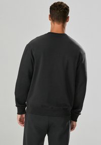 Man in een effen zwart sweatshirt en zwarte broek, gezien vanaf de achterkant tegen een neutrale grijze achtergrond.