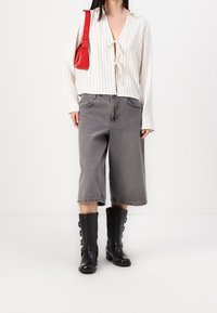 Femme portant un haut blanc à rayures fines avec liens à l'avant, un jean gris à jambes larges et coupé au-dessus de la cheville, des bottes mi-mollet noires à boucle, et portant un sac à épaule rouge.