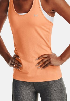 Sportowiec w brzoskwiniowej, bezrękawnikowej koszulce z logo Under Armour i białym sportowym biustonoszu, stojący z rękami na biodrach, widoczne szare legginsy.