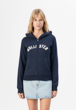 Blonde woman wearing navy zip-up hoodie with "HOLLISTER 1922" text and light blue denim skirt standing against white background.
