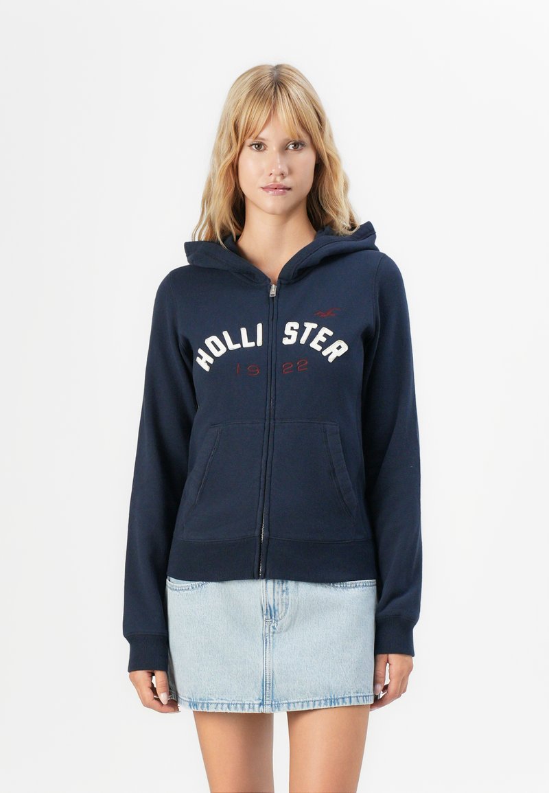 Blonde Frau trägt eine marineblaue Kapuzenjacke mit dem Aufdruck "HOLLISTER 1922" und einen hellblauen Denim-Rock, während sie vor einem weißen Hintergrund steht.
