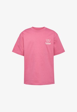 Ung pige med langt krøllet hår iført en pink hummel t-shirt, lilla joggingbukser og mørkelilla sneakers, står med hænderne i lommerne.