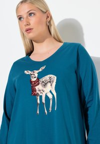 Blonde Frau, die ein türkisfarbenes Langarmshirt mit einem Aufdruck eines Rehkitzes trägt, das einen roten Pailletten-Schal umhat.