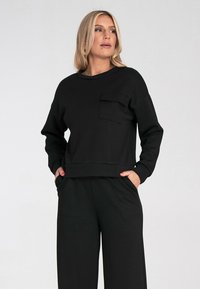 Czarny sweatshirt z długimi rękawami, okrągłym dekoltem i przednią kieszenią. Miękka tkanina i luźny krój. Połączony z dopasowanymi czarnymi spodniami.