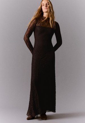 Femme aux longs cheveux blonds portant une robe longue en dentelle noire à manches longues, debout devant un fond gris uni.