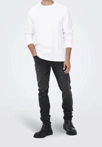 Maglietta bianca a maniche lunghe abbinata a jeans scuri e stivaletti neri. L'outfit presenta un design semplice con una vestibilità comoda e colori solidi.