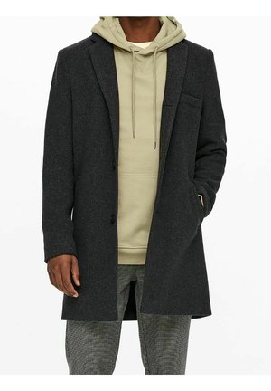 Homme portant un manteau long gris foncé par-dessus un sweat à capuche beige clair et un pantalon gris à carreaux, mains détendues sur les côtés et dans la poche.