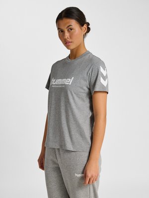 Jonge vrouw die een grijs hummel sport-T-shirt en bijpassende broek draagt, staand tegen een effen lichte achtergrond.