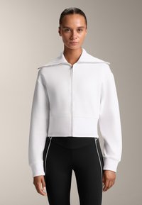 Giacca bianca corta con zip, colletto alto e polsini a costine, abbinata a leggings neri con accenti bianchi sui lati.