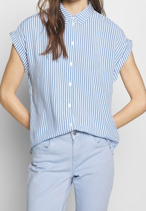 Femme portant une chemise à rayures verticales bleues et blanches à manches courtes boutonnée, rentrée dans un pantalon bleu clair, mains dans le dos.