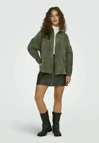 Jaqueta de camisa em corduroy verde-oliva com bolsos frontais, usada sobre um suéter de gola alta branco, combinada com uma saia mini de couro preta e botas de tornozelo pretas.