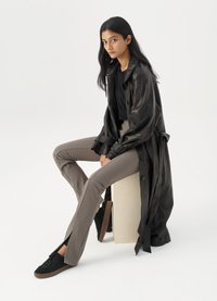 Trench coat din piele neagră, top negru ajustat, pantaloni bej ajustați și adidași negri. Modelul stă pe un cub, având părul lung și drept.