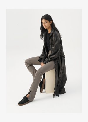 Zwarte leren trenchcoat, fitted zwart top, tan fitted broek en zwarte sneakers. Model zit op een cube, met lang steil haar.