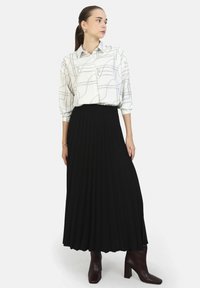 Camicetta bianca con motivo geometrico nero, abbinata a una gonna plisséc nera. Stivali marroni con tacco a blocco completano il look.