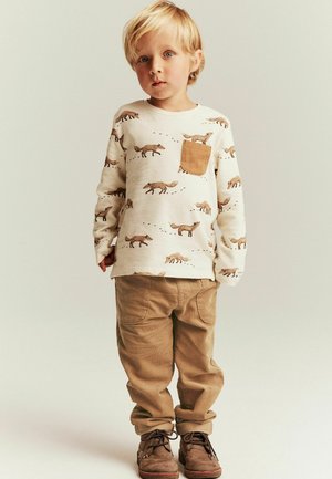 Pull clair avec des imprimés de renard et une poche marron, associé à un pantalon en velours côtelé beige et des bottes lacées marron.