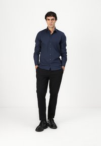 Mornarska majica dugih rukava s gumbima sprijeda, uparen s crnim hlačama i crnim cipelama. Majica ima slim fit kroj i glatku teksturu.