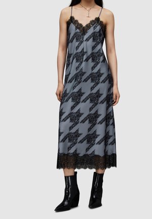 Vrouw draagt een grijze slipjurk met zwarte kantafwerking en een groot zwart ruitjespatroon, gecombineerd met zwarte puntige enkellaarsjes.