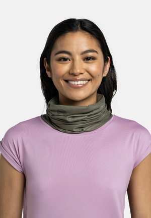 Jeune femme aux longs cheveux foncés portant une chemise violet clair et un tour de cou vert olive, souriant sur un fond blanc uni.