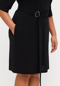 Černé šaty s krátkým rukávem, páskované v pase stříbrnou kruhovou sponou. Mají boční kapsy a hladkou, strukturovanou látku.