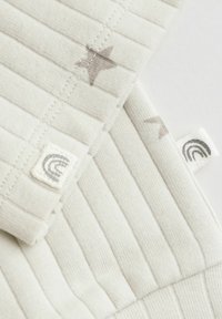 Lys cremet ribbet stof med diskrete grå stjernemønstre. Har påsyede mærkater, et af dem med et regnbuedesign på en tekstureret baggrund.