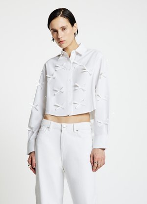 Chemisier blanc court avec un devant boutonné, orné de petits nœuds blancs. Associé à un pantalon blanc taille haute. Tissu lisse.