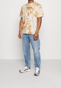 Muž v oranžovém a bílém tričku s tie-dye Kani, světle modrých džínách a bílých, stříbrných a zlatých sportovních botách stojící na neutrálním pozadí.