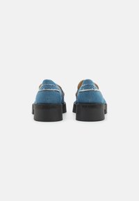 Et par blå denim slip-on loafers med sorte chunky såler, synlige sting og "Steve Madden" branding på bagsiden af hælene.