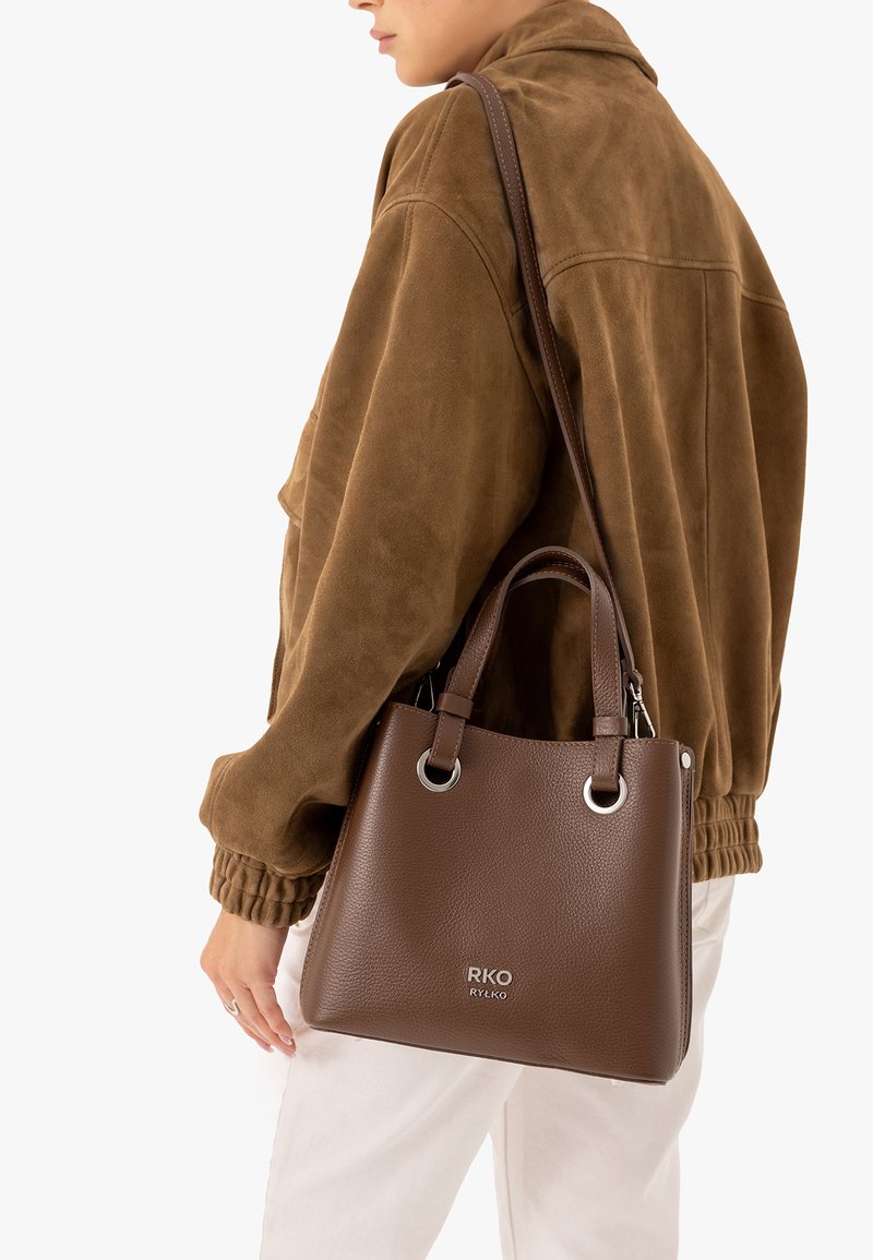 Personne portant une veste marron et un pantalon blanc porte un sac à main en cuir marron avec des anneaux argentés et le logo RKO RYŁKO à l'avant.