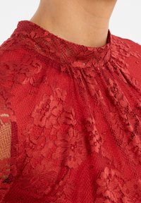 Rode kanten top met een hoge halslijn, voorzien van bloemenpatronen, gestructureerde stof en subtiele gathers bij de kraag.