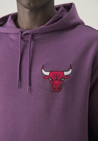 Sudadera morada con un logo de toro rojo bordado en el pecho, con un bolsillo frontal y capucha con cordón. El tejido parece suave y liso.