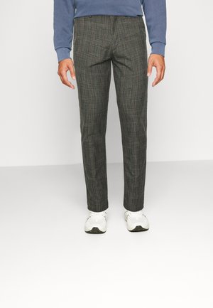 Homme portant un pantalon slim à carreaux gris, des baskets blanches et un haut bleu à manches longues, debout devant un fond uni.
