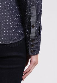 Chemise bleu marine à pois avec un poignet à boutons, présentant une texture douce et des pois bleu clair contrastants. Un denim foncé est porté en dessous.