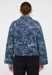 Veste en denim bleu offrant un motif texturé, des poignets larges et un col classique. Le design présente des formes abstraites et des nuances variées.