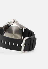 Montre avec bracelet en caoutchouc noir et boîtier en acier inoxydable, présentant un dos texturé et une boucle en argent. Comprend plusieurs trous d'ajustement pour le bracelet.