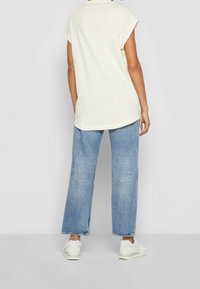 Krémové bavlněné tričko s ohrnutými krátkými rukávy, spojené se světle modrými širokými džíny a bílými teniskami, viděné zezadu.