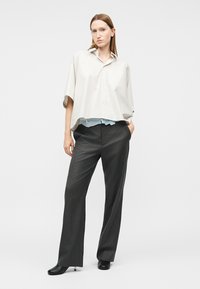 Naine seisab käed taskutes, tal on seljas lahtine valge lühikeste varrukatega särk, tumedad laiad püksid ja mustad kontsakingad ühtlasel taustal.