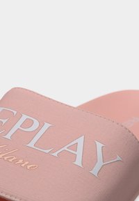 Ροζ σανδάλι με φαρδιά λωρίδα από υφασμάτινη υφή και λευκό κείμενο λογότυπου. Η σόλα είναι ελαφρώς καμπυλωτή και λεία.