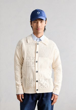 Ung mand iført en cremefarvet strikket cardigan med knapper og teksturerede mønstre, en blå kasket med hvid globuslogo og mørkeblå jeans.
