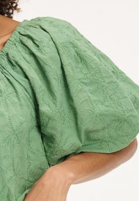 Blusa verde ricamata con un motivo floreale testurizzato, maniche a sbuffo e scollatura arricciata. Tessuto leggero con una finitura morbida.