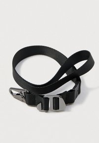 Sangle en nylon noir avec une texture lisse, dotée d'une boucle et d'un fermoir en argent. La sangle est plate et enroulée dans une position défaite.