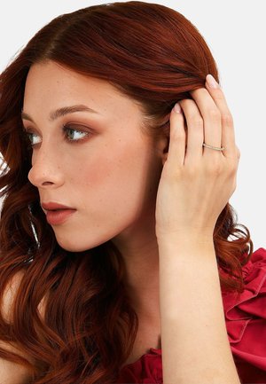 Junge Frau mit roten Haaren, hinter dem Ohr gesteckt, trägt einen zarten Diamantring und ein rotes gerüschtes Oberteil, schaut nach links.