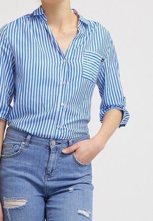 Persona con una camisa de botones a rayas verticales azules y blancas con las mangas remangadas y jeans azules desgastados, con la mano en el bolsillo.