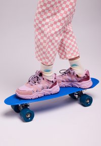 Un pantalon à motifs à damier rose posé au-dessus d'un skateboard bleu, avec des baskets à motifs rose et violet avec des accents duveteux et des rayures noires.