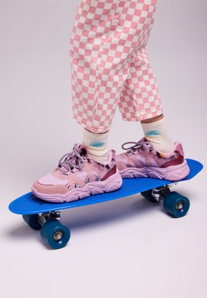 Un pantalon à motifs à damier rose posé au-dessus d'un skateboard bleu, avec des baskets à motifs rose et violet avec des accents duveteux et des rayures noires.