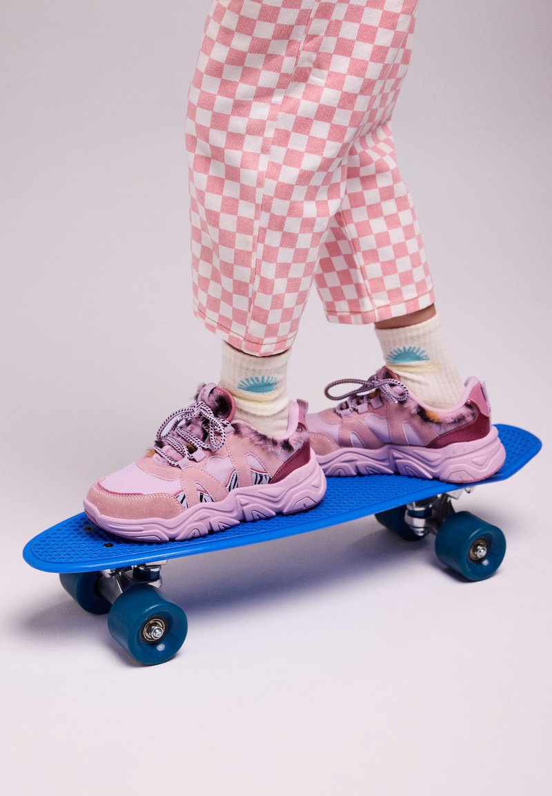 Un pantalon à motifs à damier rose posé au-dessus d'un skateboard bleu, avec des baskets à motifs rose et violet avec des accents duveteux et des rayures noires.
