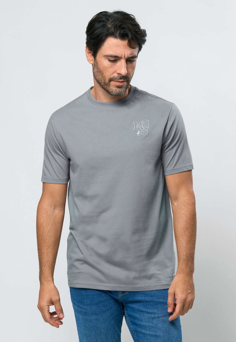 Homme aux cheveux noirs et barbe portant un t-shirt gris clair avec un logo blanc sur la poitrine et un jean bleu, regardant vers le bas avec une expression neutre.