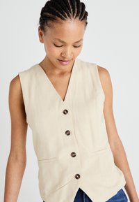 Beige ärmellose Weste mit V-Ausschnitt, ausgestattet mit vier dunklen Knöpfen und zwei seitlichen Taschen, aus einem glatten Stoff mit einer dezenten Textur.